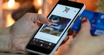 Eine Frau öffnet auf ihrem Smartphone einen Online-Shop. In der rechten Hand hält sie ihre Kreditkarte für die Zahlung bereit.