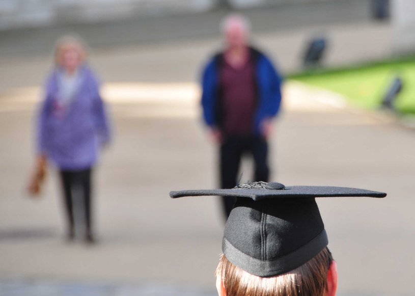 Die Rückansicht eines Studierenden. Auf dem Kopf trägt er einen Bachelor-Hut. Im Hintergrund sind zwei Personen auf einem Platz zu sehen.