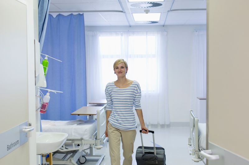 Eine Frau verlässt glücklich ein Krankenhauszimmer mit ihrem Rollkoffer in der Hand.