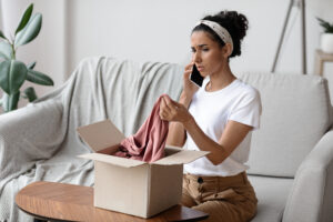 Disappointed young brunette woman sitting on sofa by wooden table and opening carton box, talking on mobile phone with seller after checking delivery, holding new clothes and complaining to customer service.