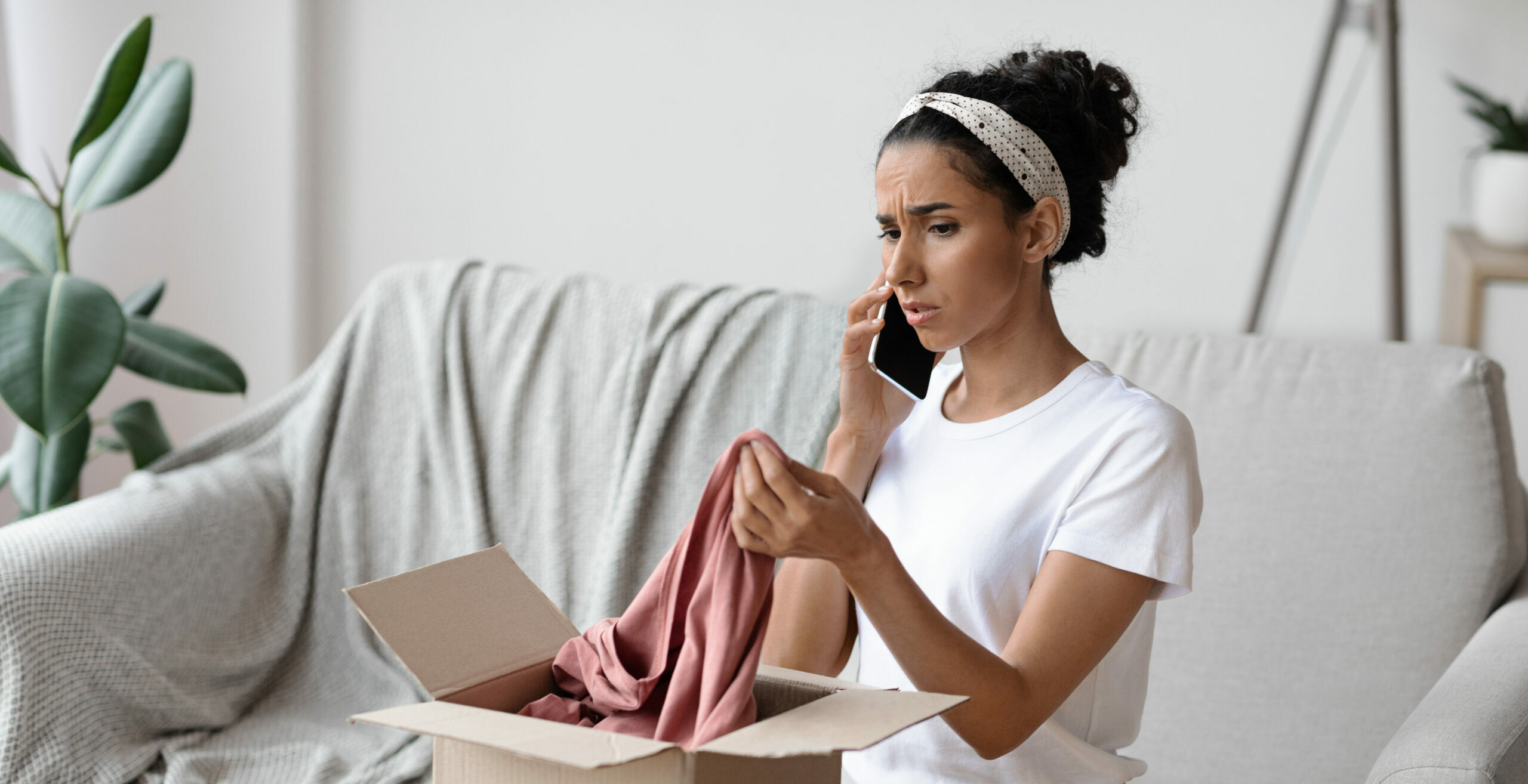 Enttäuschte junge brünette Frau sitzt auf dem Sofa an einem Holztisch und öffnet einen Karton. Sie telefoniert mit dem Verkäufer, nachdem sie die Lieferung überprüft hat, hält neue Kleidung in der Hand und beschwert sich.