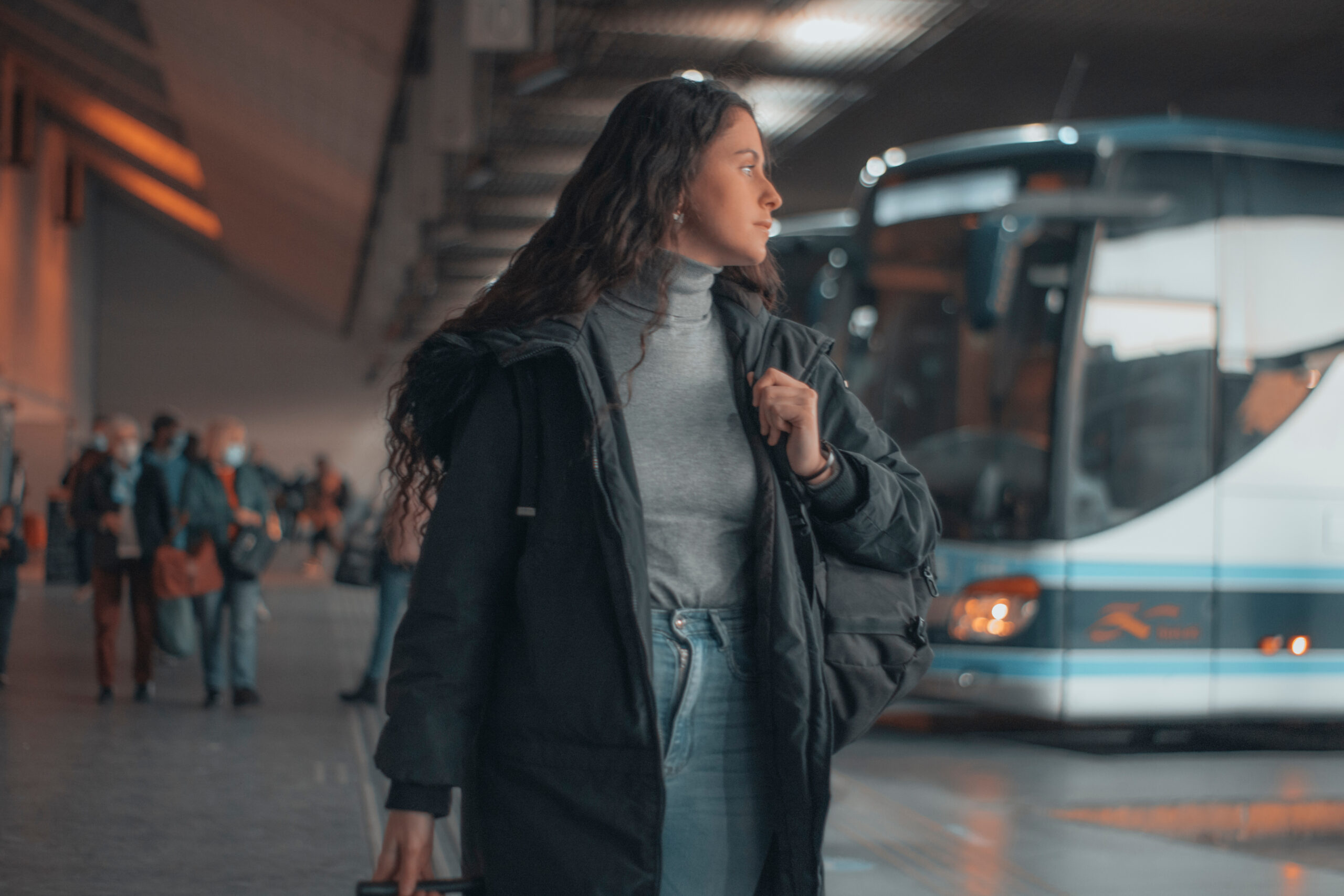 Dunkelhaarige Reisende, die ungeduldig und mit Rucksack und Reisekoffer am Busbahnhof wartet.