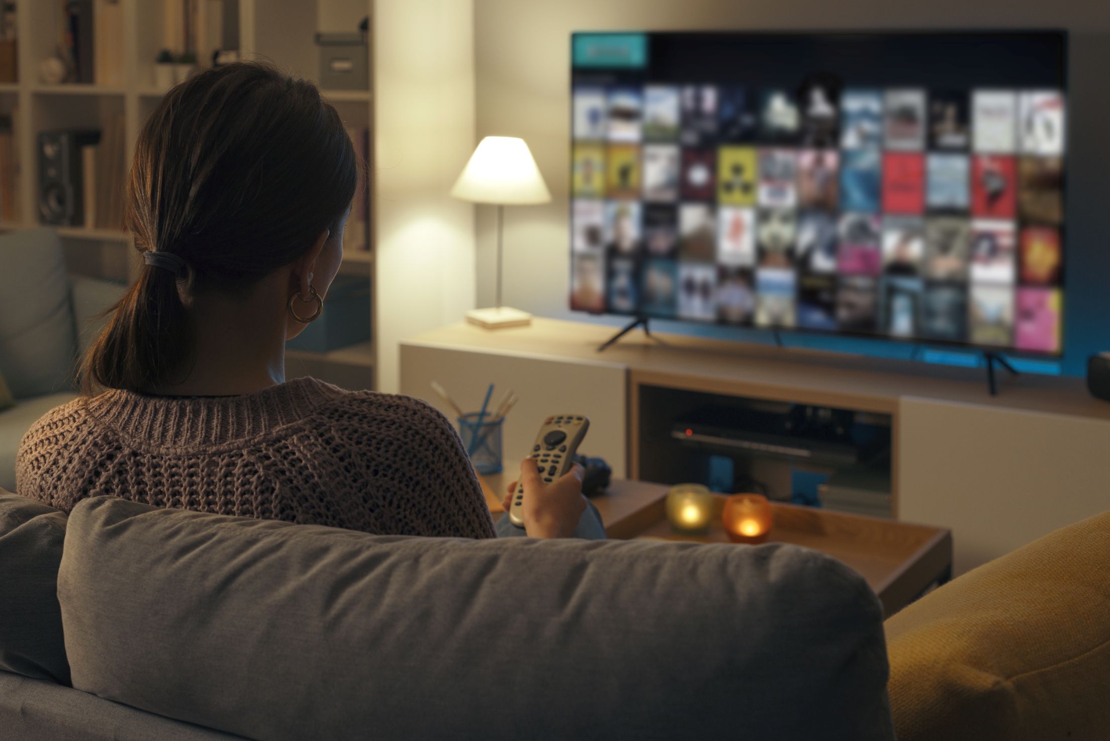 Eine Frau sitzt auf der Couch und klickt sich mit der Fernbedienung durch das Angebot eines Streaming-Portals.