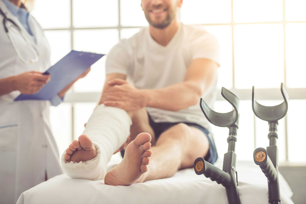 A patient sits on a lounger. He has a plaster on his right foot. An illustration of an article on emergency treatment in Germany.