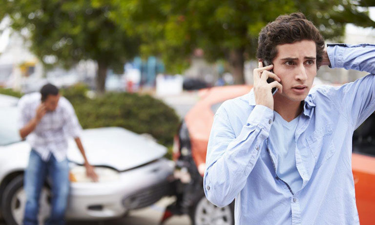 In the foreground is a young man on the phone. He has his left hand on the back of his head and his smartphone in his right hand. In the background, there is a car crash.