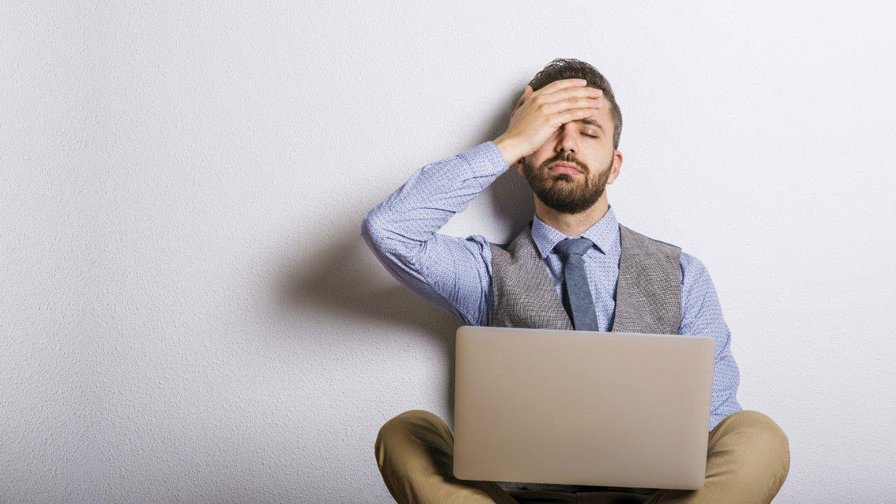 A man sits in front of his laptop and holds his hand to his head.