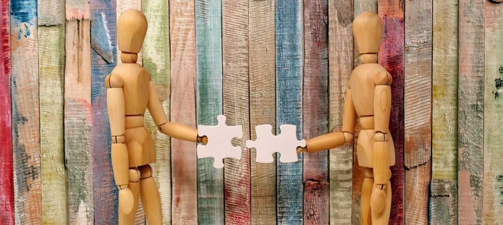 Zwei Holzfiguren strecken sich ein Puzzleteil zu