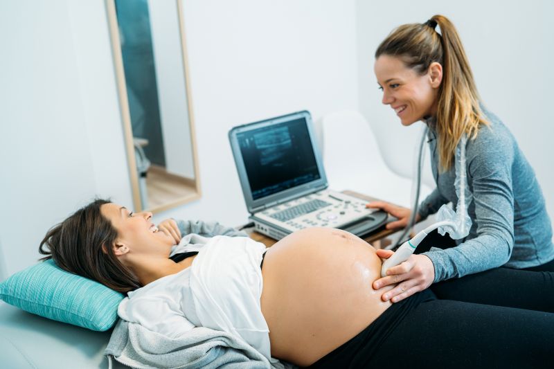 Eine hoch schwangere Frau spricht während einer Ultraschalluntersuchung fröhlich mit der Ärztin.