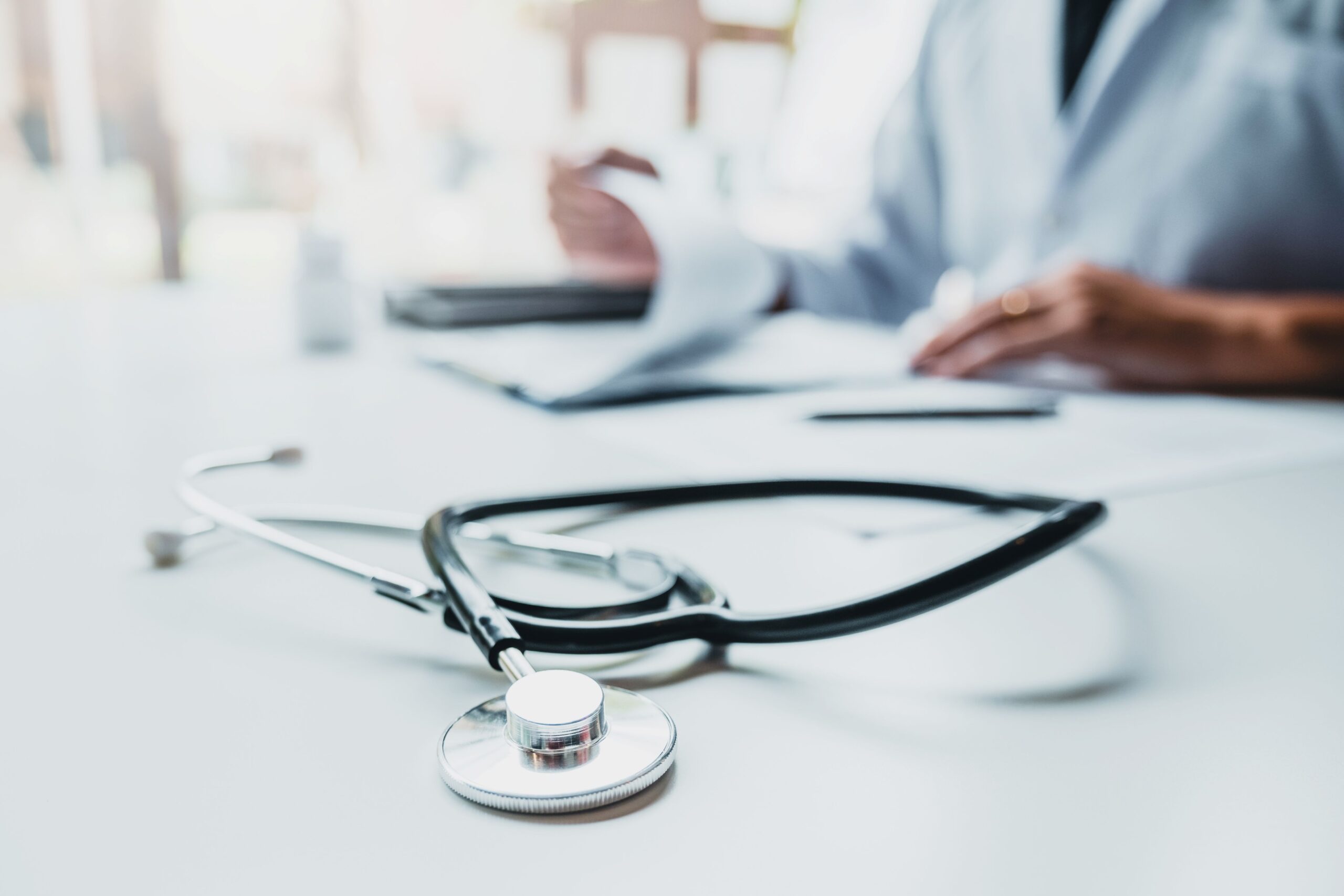 A stethoscope is lying on the table. In the background, a doctor is going through some documents.