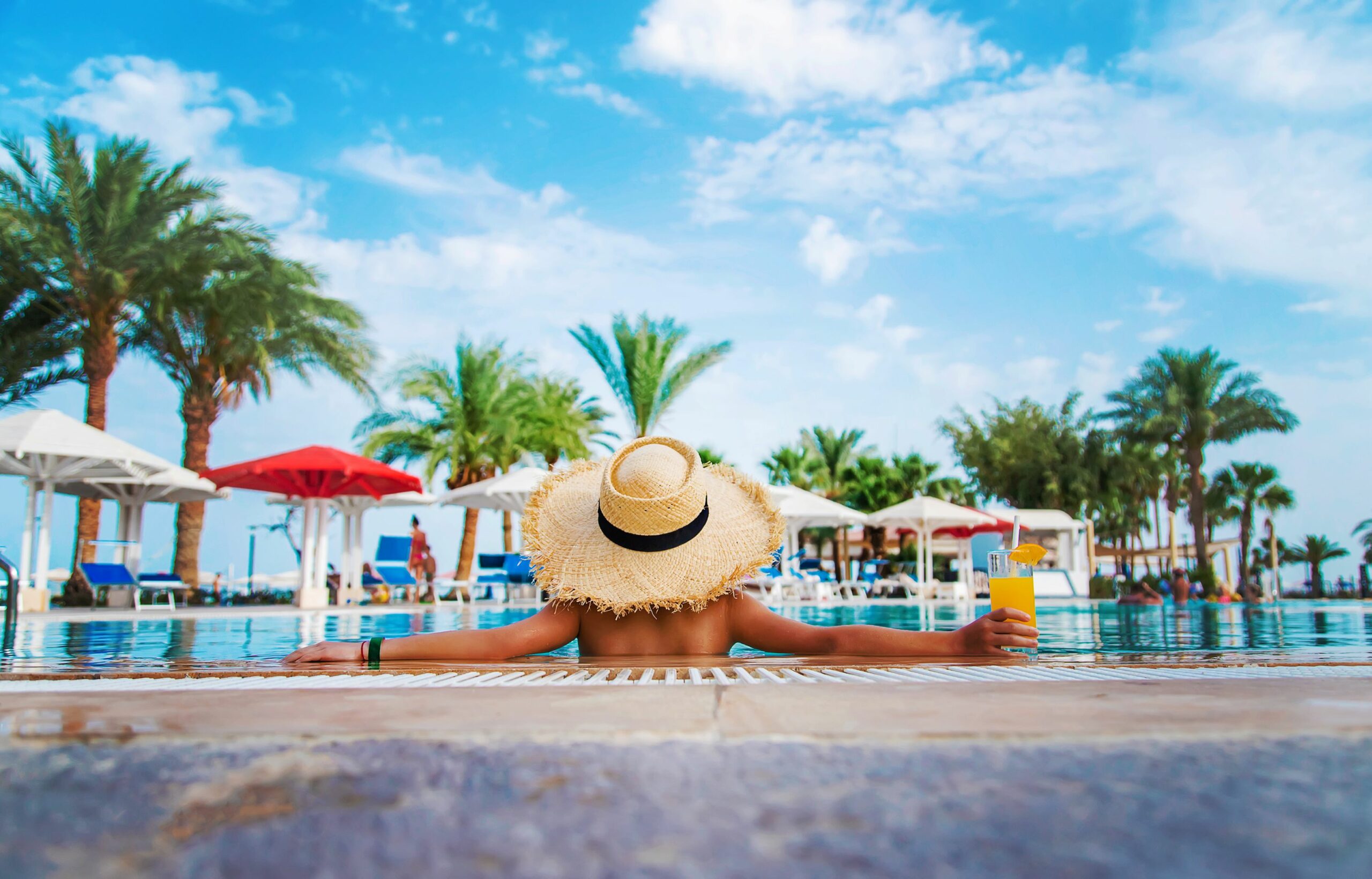 A person wearing a straw hat is sitting in the pool with a glass of fruit juice in their hand. Palm trees can be seen in the background.