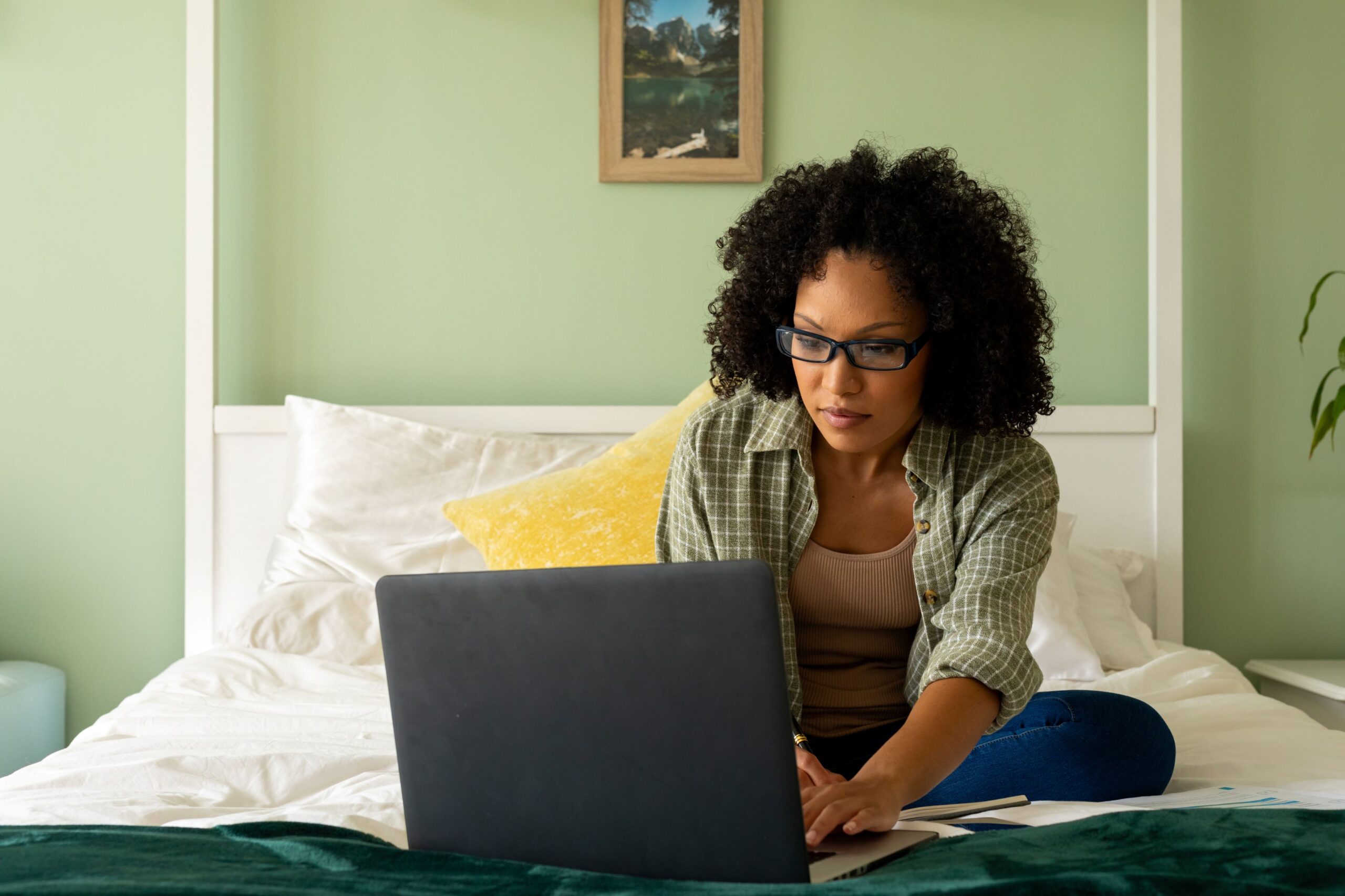 A woman is sitting on her bed, looking for something on her laptop.