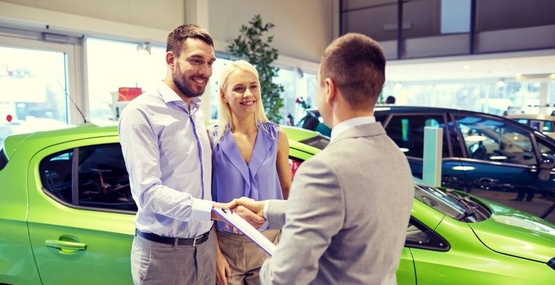 A couple is standing in front of a green car with a salesperson at the car dealership.