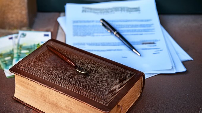 Ein Faksimile, ein braunes Lederbuch, Hunder-Euro-Scheine und Unterlagen auf einem Tisch