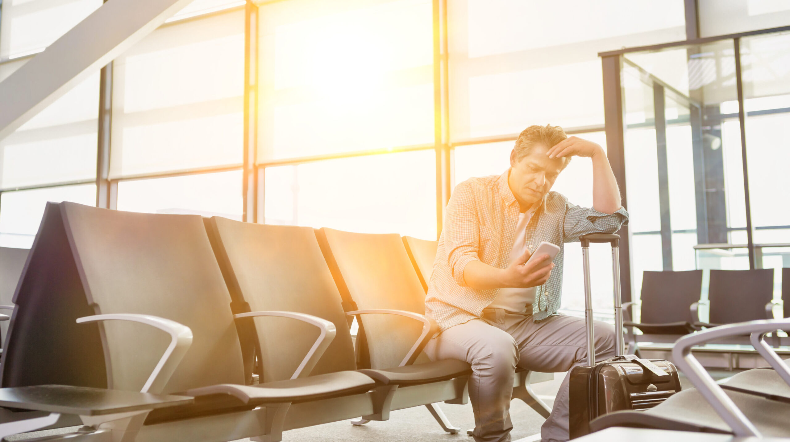 A stressed man sits on a bench in an airport terminal, staring at his mobile phone