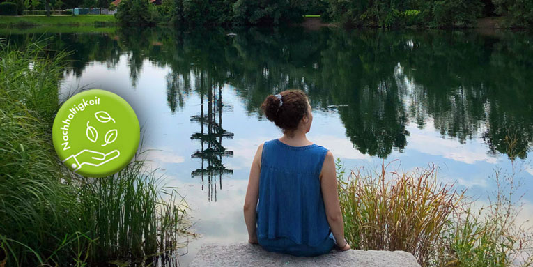 Eine junge Frau mit blauem Oberteil sitzt auf einem Stein an einem See.