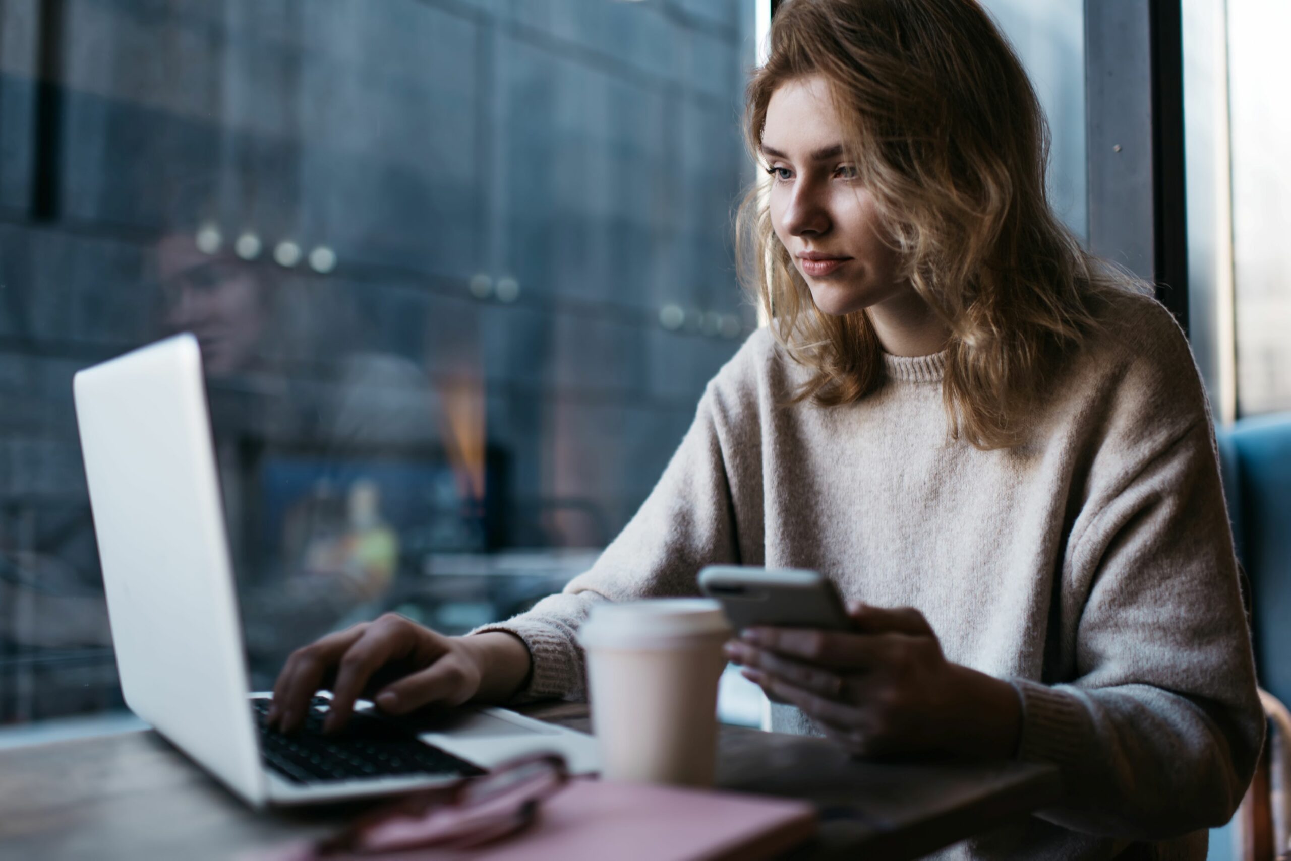 A woman is sitting in a café working on a laptop and holding her smartphone in her hand.