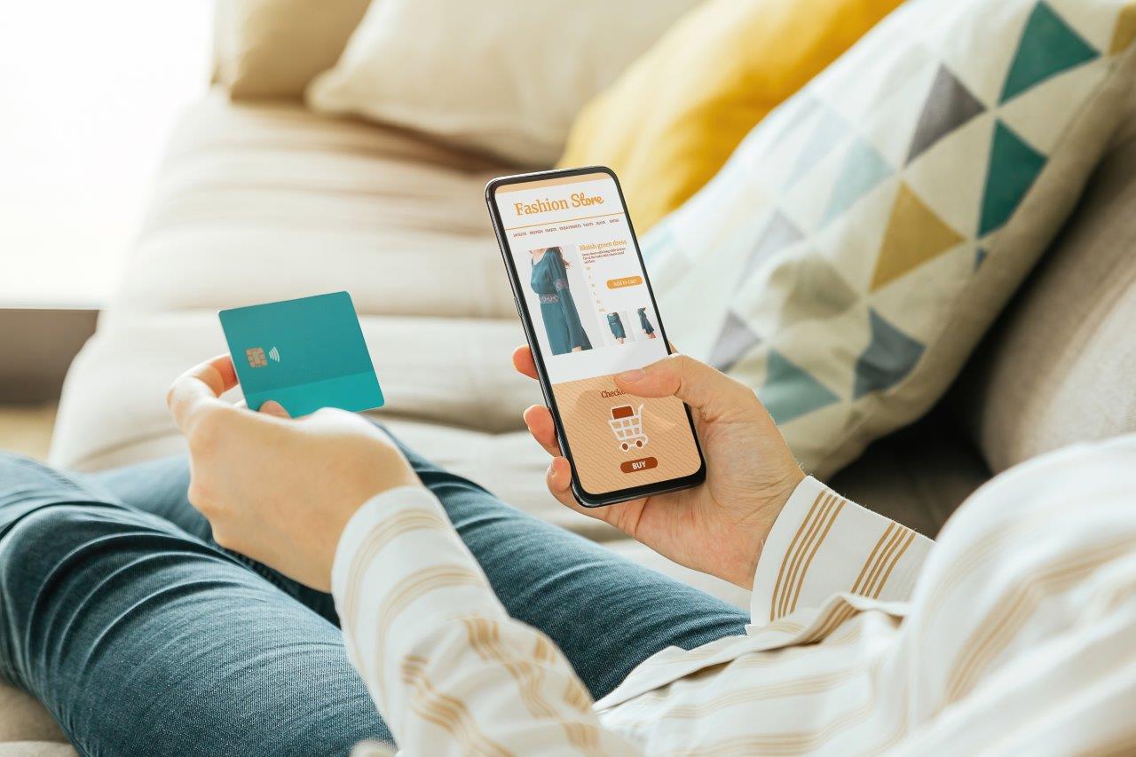 Eine Person beim Online-Shopping: In der rechten Hand hält sie das Smartphone, auf dem die Webseite mit dem Warenkorb angezeigt wird. In der linken Hand hält sie die Kreditkarte