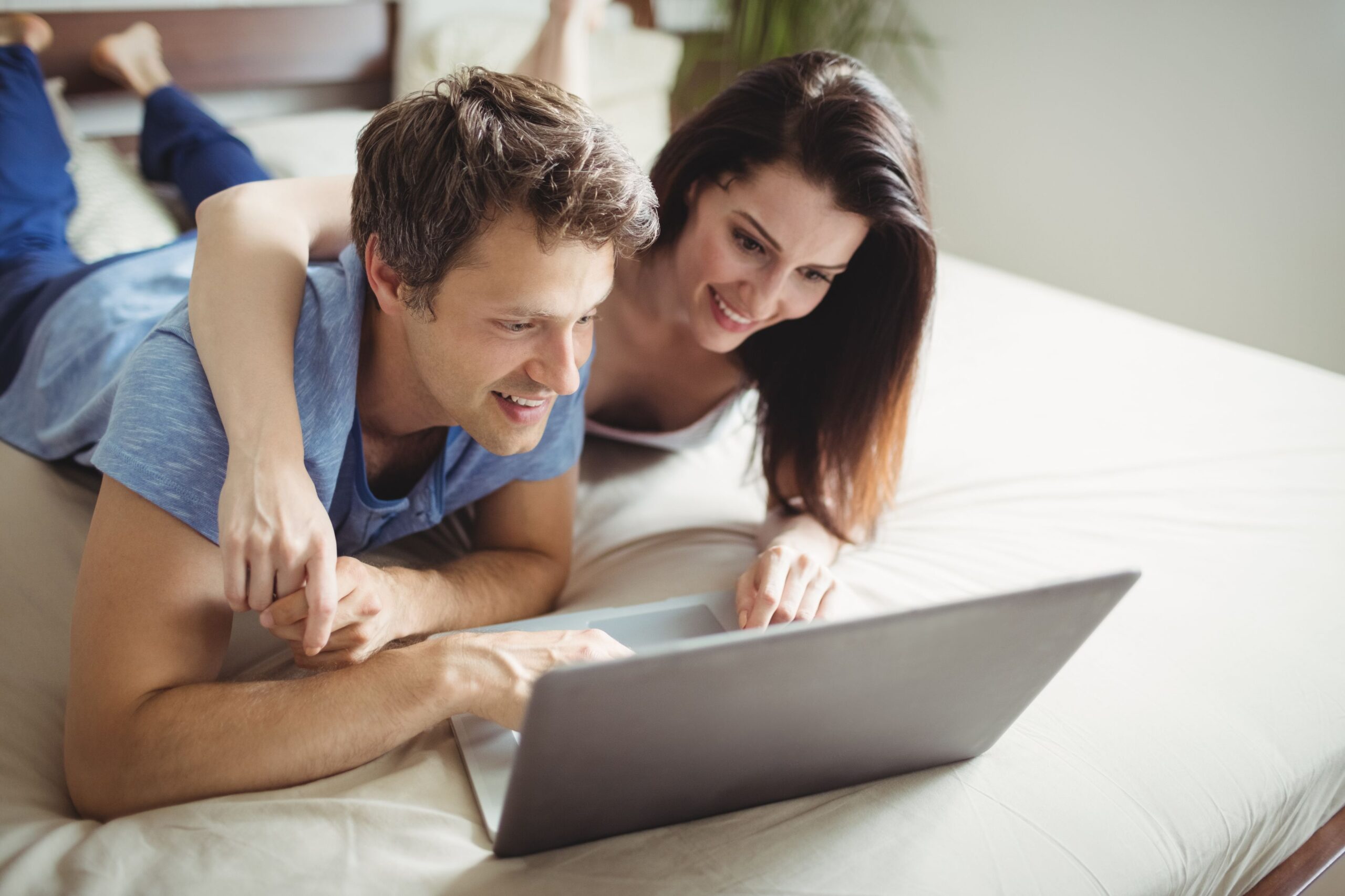 A couple are lying on the bed, looking something up together on a laptop.