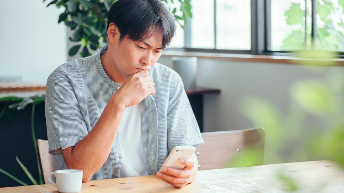 Ein Mann sitzt in einem Café am Tisch und schaut nachdenklich auf den Bildschirm seines Smartphones. Seine Stirn ist gerunzelt, die Hand am Kinn.