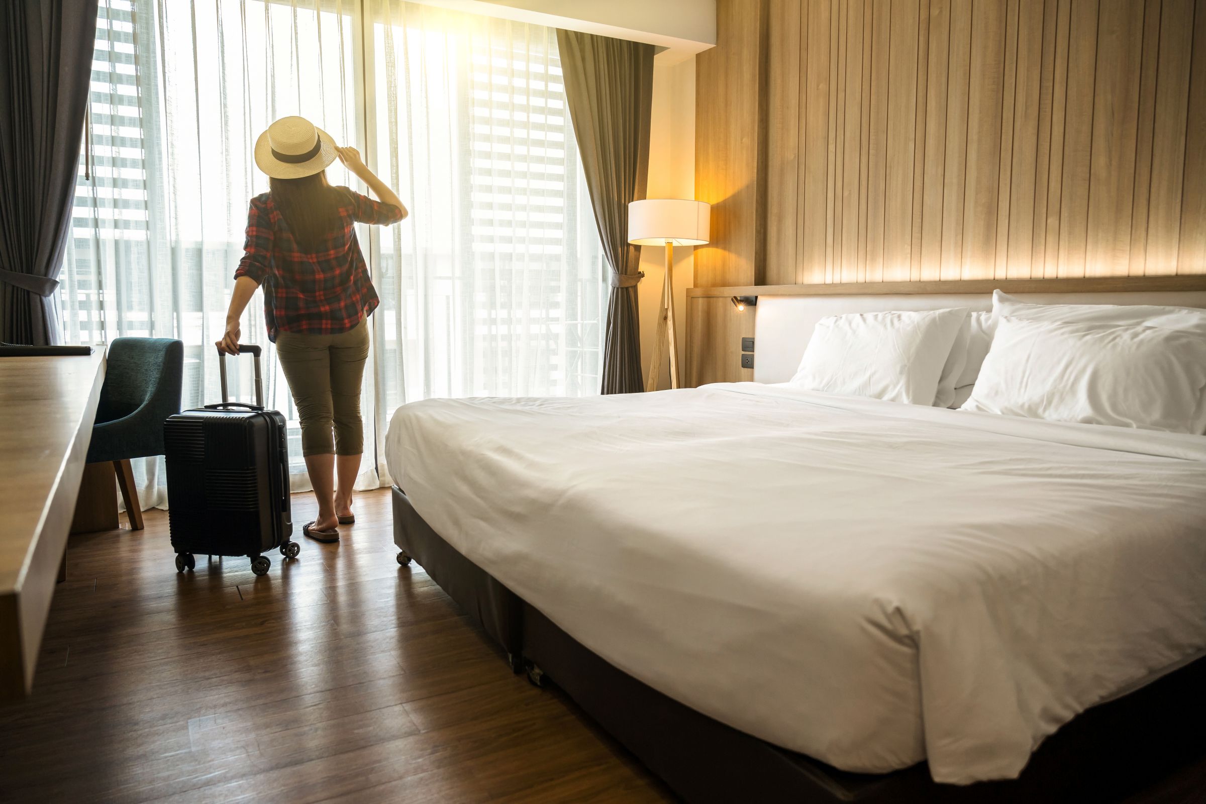 Eine Frau steht mit ihrem Koffer im Hotelzimmer am Fenster und blickt hinaus.