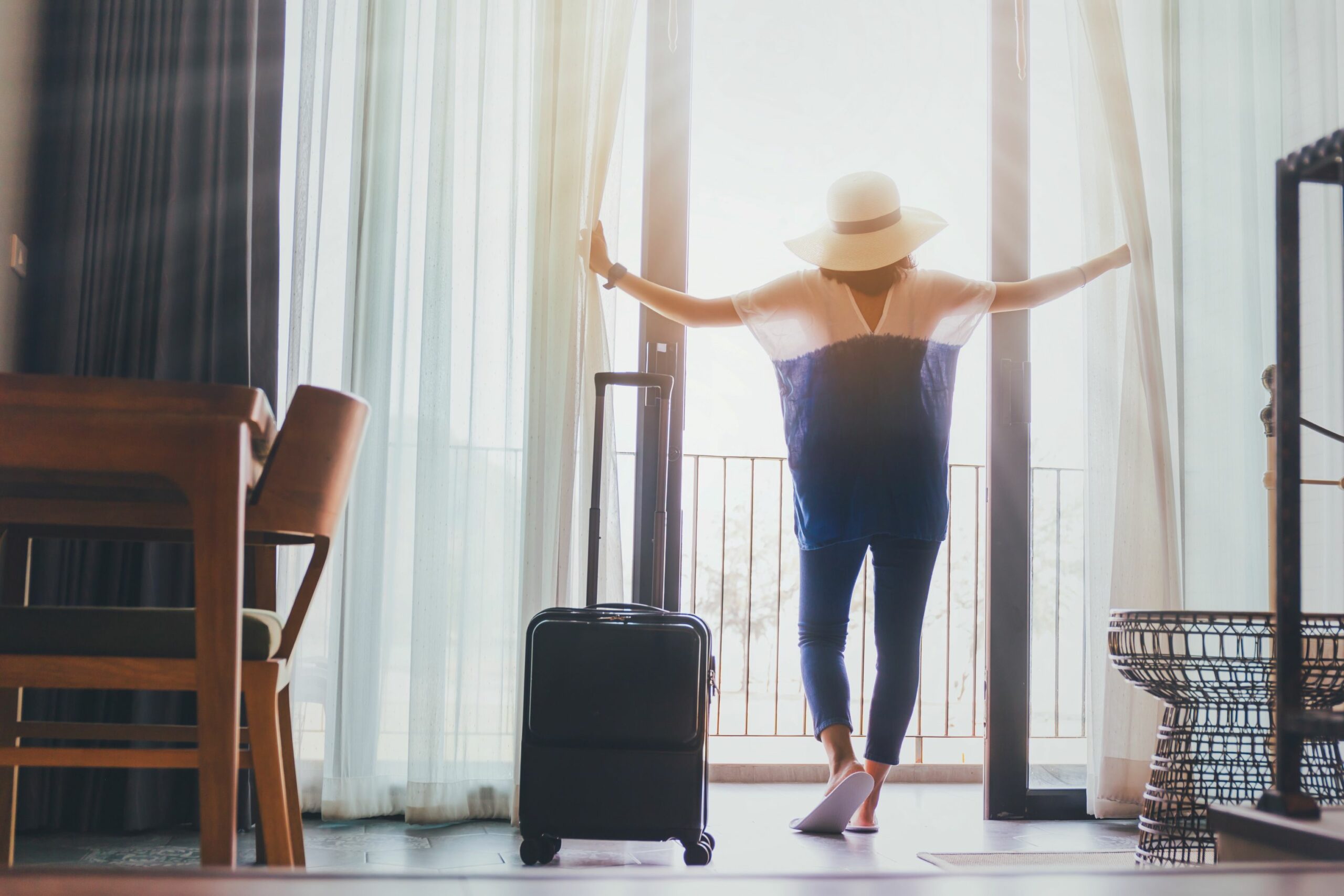 Eine Frau steht vor dem geöffneten Fenster des Hotelzimmers und blickt hinaus. Links von ihr steht ein Koffer.