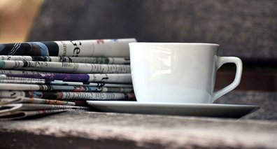 Ein Zeitungsstapel liegt auf einer Holzbank. Daneben steht eine weiße Kaffee-Tasse.
