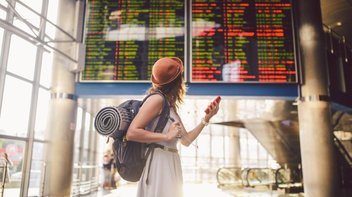 Frau steht mit Handy im Flughafenterminal und schaut auf Anzeigentafel 