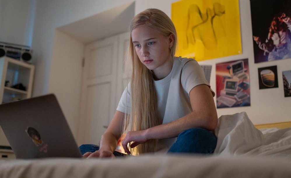 A teenager girl is sitting on a bed and uses a laptop.