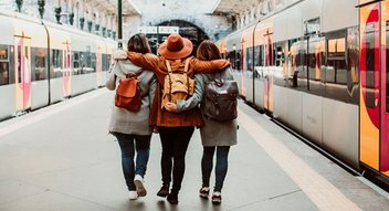 Drei Personen umarmen sich am Bahnsteig. Links und rechts stehen Züge.
