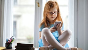 Ein kleines Mädchen mit roten Harren und Brille sitzt im Schneidersitz und spielt mit einem Smartphone.