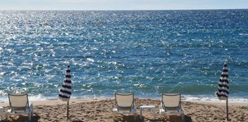Drei weiße Liegestühle stehen am Strand mit Blick auf das Meer.