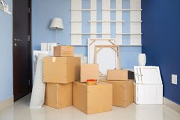 Moving boxes, rolled carpet and other objects in front of a blue corridor wall.