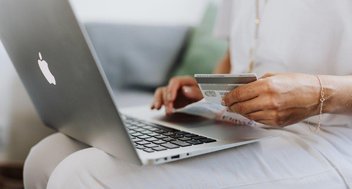 Eine Frau hat auf ihrem Schoß einen Laptop stehen. In der Hand hält sie eine Kreditkarte.