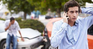 A young man is calling someone with his smartphone. In the background is a car accident.